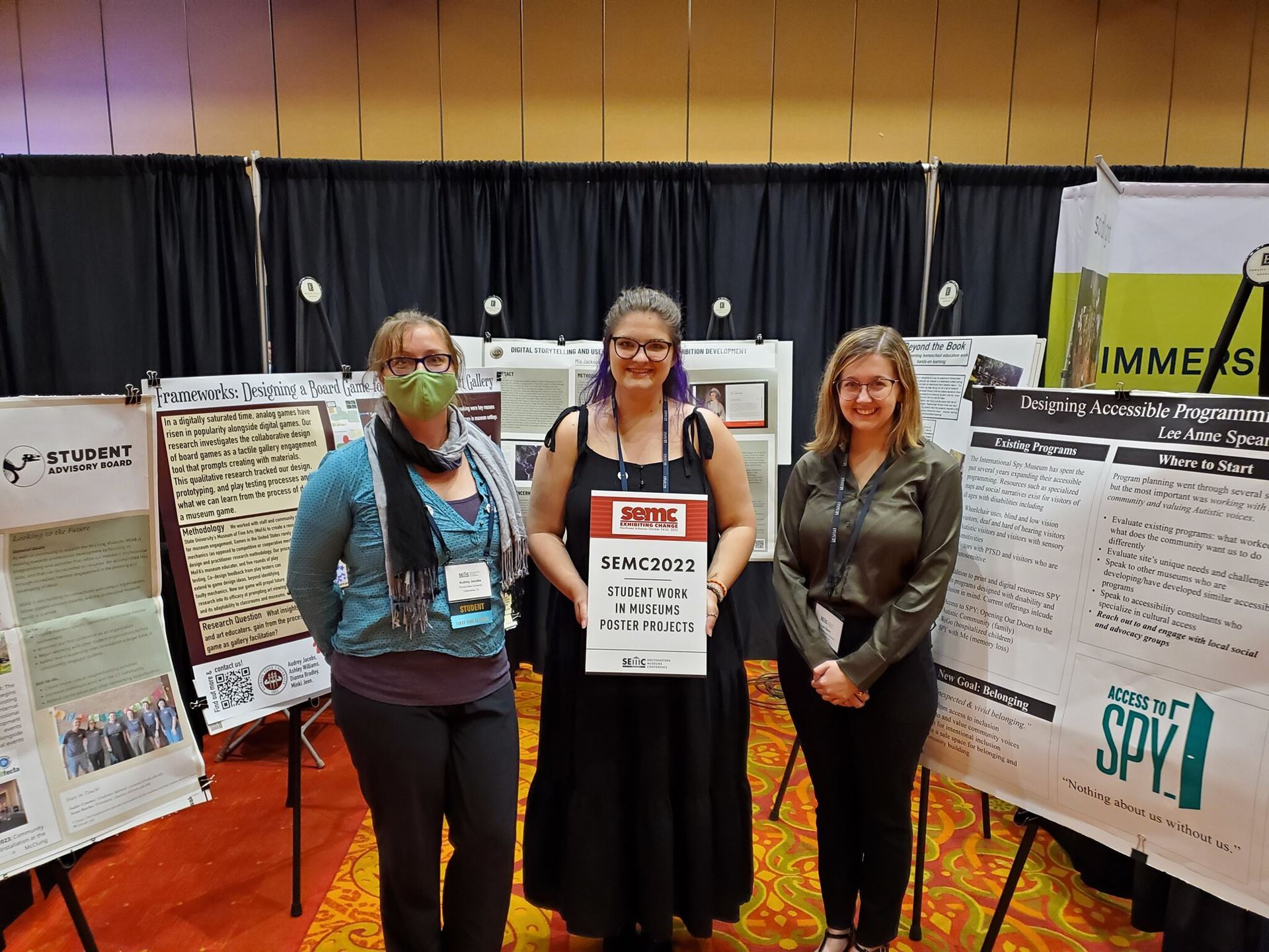 Three women stand centered in the picture, all smiling at the camera. Surrounding them are many poster presentations. The woman in the middle holds a sign that says "SEMC2022: Student Works in Museums Poster Projects"