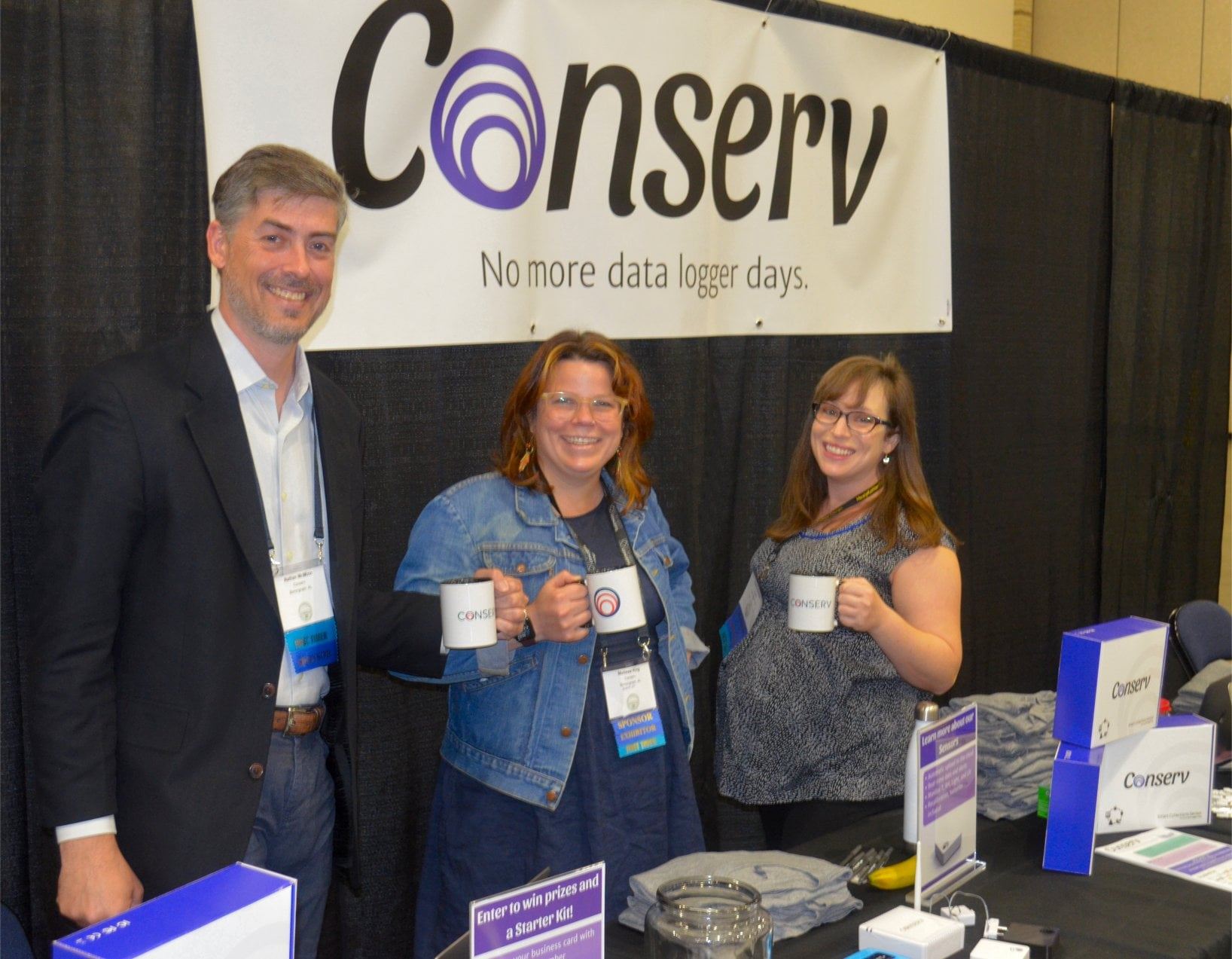 Medium shot of three Conserv team members at a stand with Conserv products. All three members are standing, smiling, and holding conserv mugs against a black curtain and a sign that reads "Conserv no more data logger days".