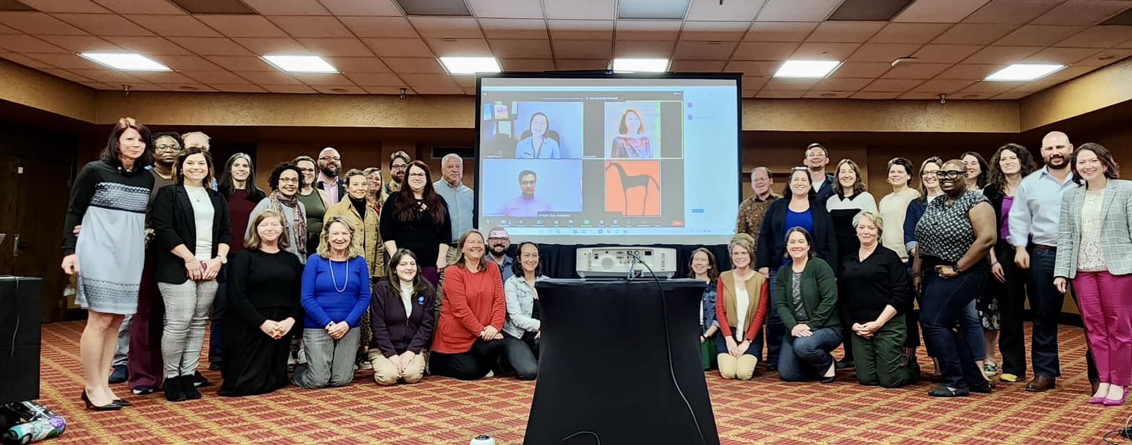 the SEMC2023 Program Committee meets both in person and on Zoom in Louisville, Kentucky. All the people pictured are smiling at the camera, and a screen showing the virtual attendees is pictured in the middle of the group.