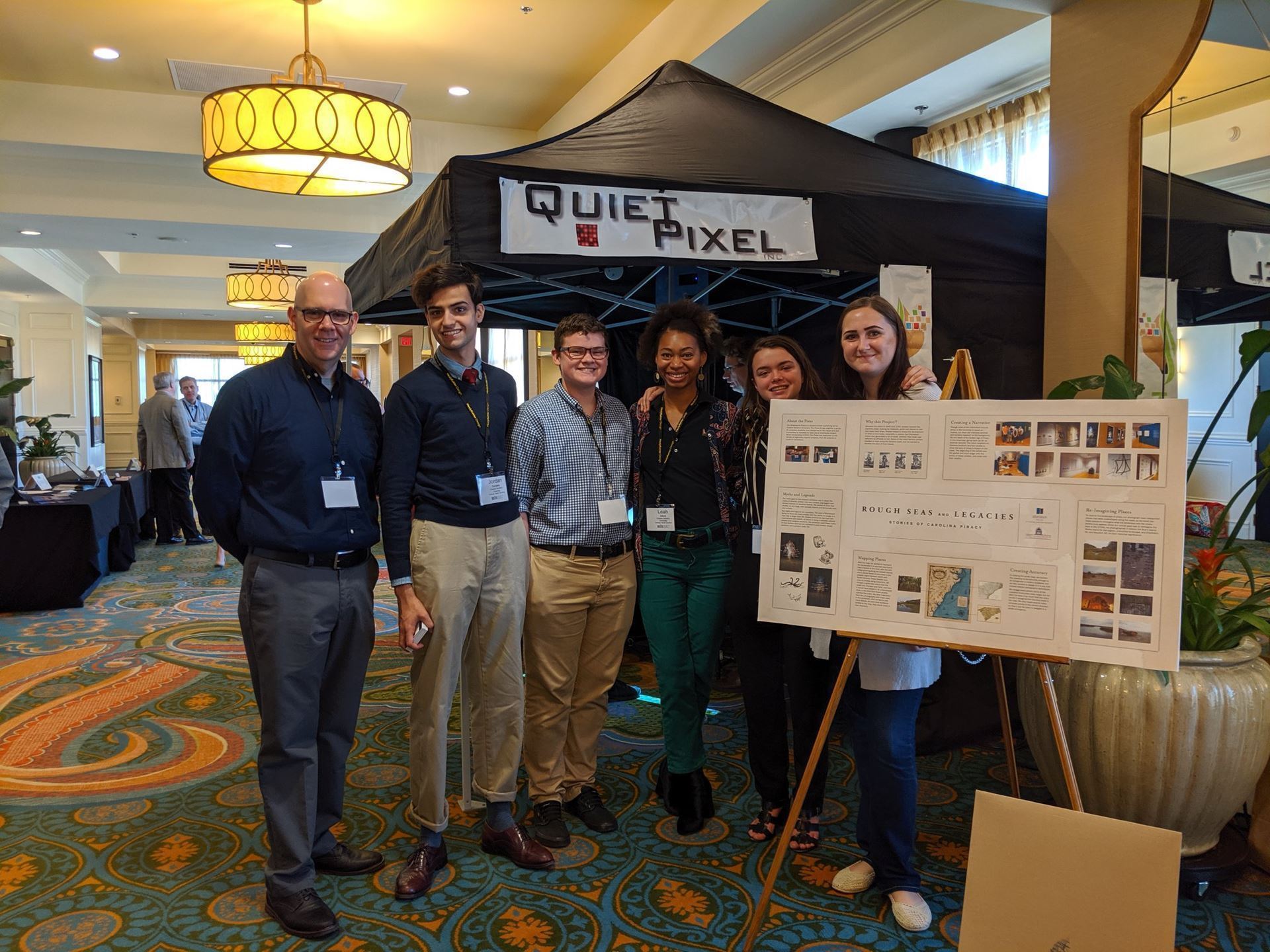 5 people stand next to each other and smile at the camera, in front of the two people on the right, there is a poster presentation on an easel. Behind them is a black tent that says "Quiet Pixel". They stand in a hallway.